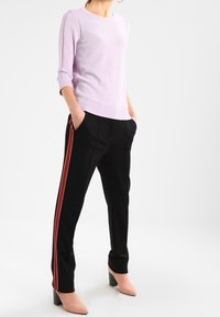 Liliowy sweter na drutach z rękawami do łokcia, w połączeniu z czarnymi spodniami z czerwono-białymi paskami po bokach i jasnoróżowymi szpilkami.