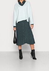 Pull bleu clair surdimensionné avec un col en V profond, porté sur une robe boutonnée vert foncé présentant un petit motif floral. Bottines noires à talons.