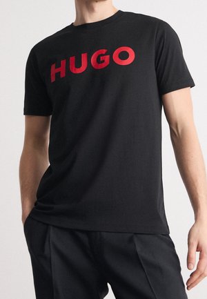 Mand iført sort kortærmet T-shirt med fed rød "HUGO" tekst på brystet og sorte bukser, stående med hænderne nær lommerne.