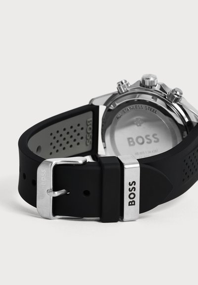 Bracelet de montre en caoutchouc noir avec boucle et fermoir métalliques argentés gravés de "BOSS,", fixé à un fond de boîtier en acier inoxydable avec "BOSS" gravé.