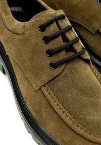 Chaussure en daim marron à lacets noirs, œillets noirs, coutures visibles et semelle noire texturée.