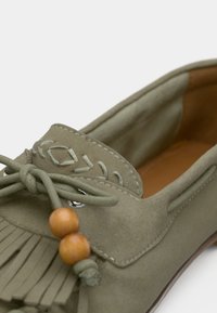 Mocassin en suède vert avec des franges, des coutures décoratives et des perles en bois. Texture douce avec un bout arrondi et un profil bas.