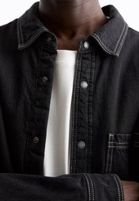 Sort denimjakke med krave, hvide syninger og trykknapper. Har en brystlomme og er lagdelt over en hvid skjorte.