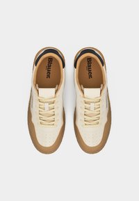 Ζευγάρι μπεζ και καφέ αθλητικά παπούτσια με κορδόνια, σόλες από καουτσούκ και μαύρες λεπτομέρειες, από πάνω σε απλό λευκό φόντο.