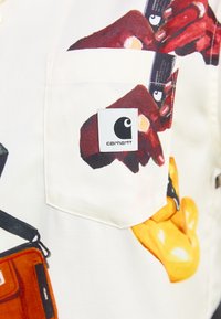 Weißes Shirt mit grafischen Prints, das Elemente wie rote Hände und ein gelbes Objekt zeigt. Beinhaltet eine Tasche mit einem Carhartt-Logoetikett.