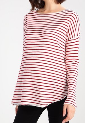 Vrouw die een wit langemouwen top met horizontale rode strepen en een zwarte broek draagt, getoond van schouders tot heupen.