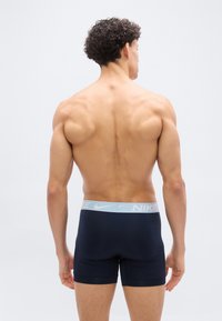 Άντρας χωρίς πουκάμισο με σγουρά μαλλιά, φοράει μαύρα εσώρουχα Nike με γαλάζια ζώνη μέσης, στέκεται γυρισμένος πλάτη σε λευκό φόντο.