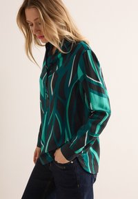 Camicia a maniche lunghe in teal e nero con motivi astratti di foglie verdi e bianche. Presenta una chiusura con bottoni e una vestibilità rilassata.