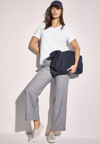 Witte V-hals t-shirt, grijze geruite wijde broek, grijze sneakers en een marineblauwe pet. Model houdt een marineblauwe sweatshirt vast en staat tegen een lichte achtergrond.