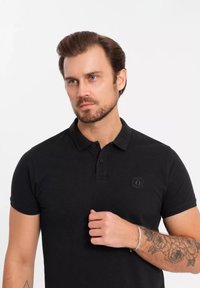 Musta polo-paita, jossa on teksturoitu kangas, taitettu kaulus, kaksinappinen etusulku ja pieni logo rinnassa. Lyhyet hihat ja kapea muotoilu.
