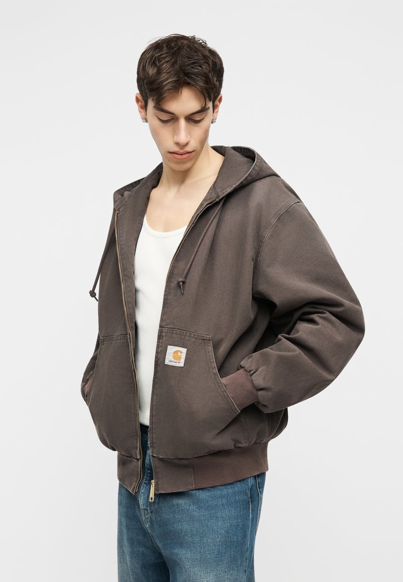 Giovane uomo che indossa una giacca con cappuccio marrone con zip sopra una canottiera bianca e jeans blu, guarda in basso con le mani nelle tasche.