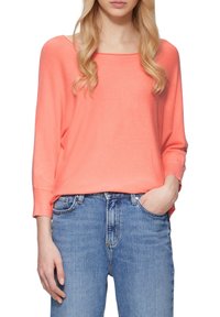 Korallenfarbener Strickpullover; lange Ärmel, weiter Ausschnitt, lockere Passform. Kombiniert mit hellblauen Jeans, die einen lässigen Look präsentieren.