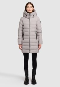 Light gray puffer coat with a high collar, front zipper, and black accents; features quilted texture and side pockets. Black boots complete the look.