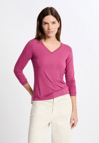 Maglietta fucsia a maniche lunghe con scollatura a V e cuciture decorative. Tessuto morbido, design aderente, con una leggera drappeggiatura in vita. Abbinata a pantaloni color crema.