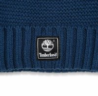 Berretto lavorato a maglia di colore blu scuro con risvolto a coste. Presenta una patch quadrata nera con un logo dell'albero bianco e la scritta "Timberland" sottostante.