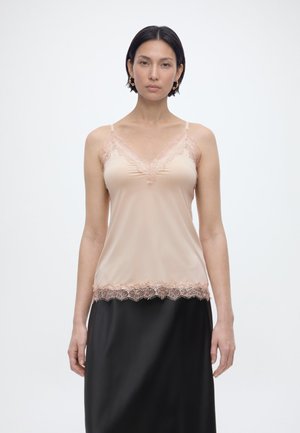 Kobieta ubrana w beżową koszulkę na ramiączkach z koronką i czarną spódnicę, stojąca na tle jednolitego białego tła.