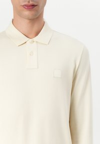 Polo à manches longues en tissu crème clair. Il présente un col polo, deux boutons et un écusson carré sur la zone de la poitrine.
