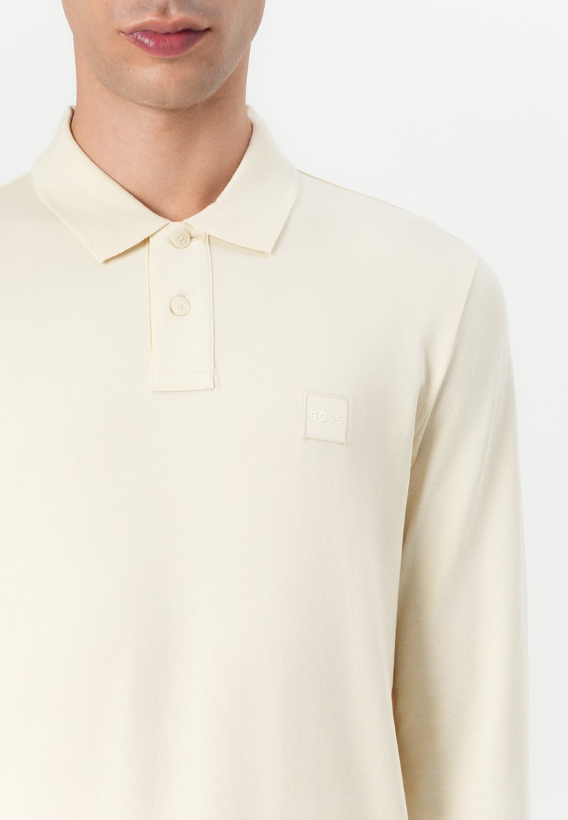 Langärmliges Polo-Shirt aus hellcremigem Stoff. Es verfügt über einen Polo-Kragen, zwei Knöpfe und ein quadratisches Logo-Label im Brustbereich.