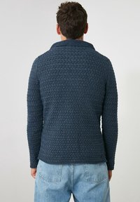 Marinblå stickad tröja med ett texturerat mönster, sjalkrage och långa ärmar, ihop med ljusblå denimjeans.