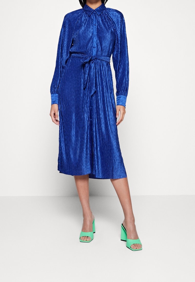 Femme portant une robe midi bleue texturée à manches longues avec ceinture à la taille et des sandales vertes à talons hauts ouvertes à l’avant, debout sur un sol blanc.