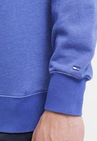 Sudadera de manga larga azul con puños acanalados y pequeño logo rectangular blanco y azul marino cerca de la muñeca, llevada en el brazo de una persona.
