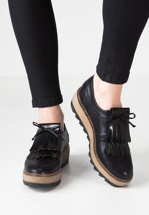 Sorte læder platformsko med tykke beige såler og frynse detaljer, båret med sorte tætsiddende bukser på et lyst gulv.