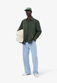 Camicia verde a maniche lunghe, jeans azzurri larghi, sneakers bianche e un cappellino verde a fantasia. Porta una borsa color crema.