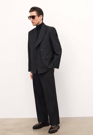 Blazer negro de doble botonadura con botones grandes, combinado con pantalones negros holgados y zapatos oscuros. Modelo llevando gafas de sol tintadas.