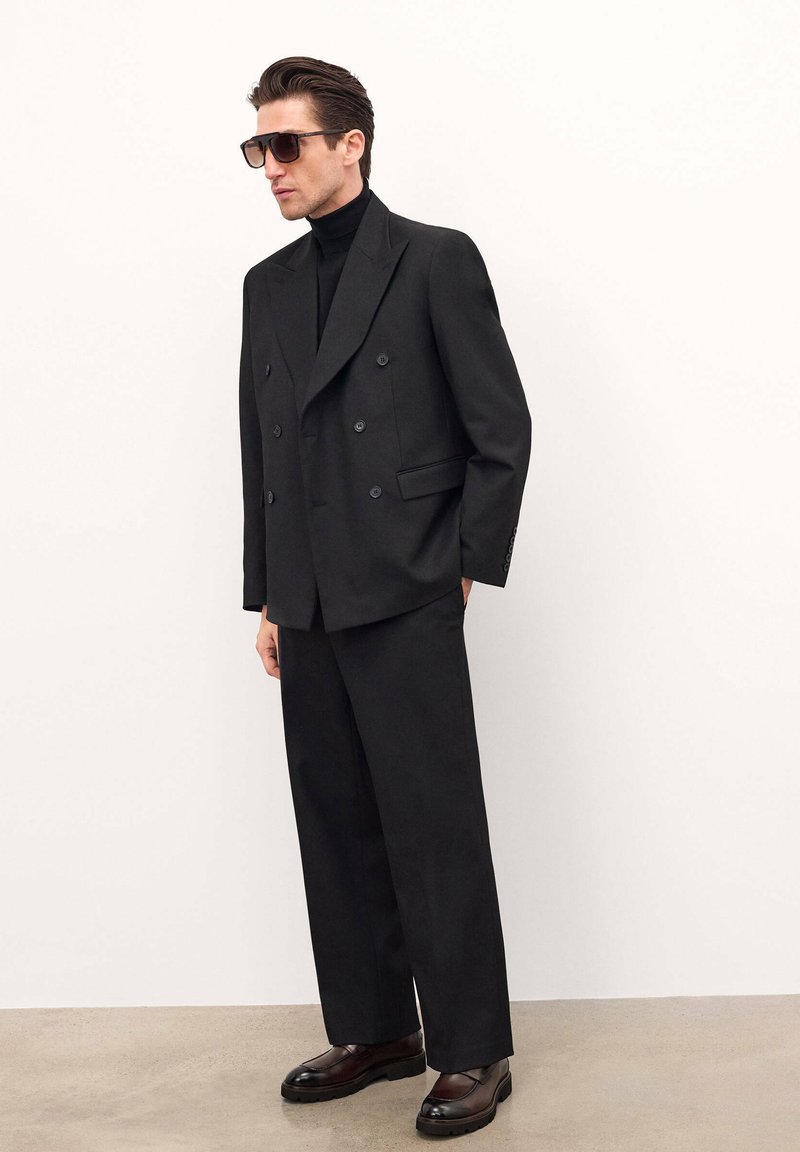 Blazer negro de doble botonadura con botones grandes, combinado con pantalones negros holgados y zapatos oscuros. Modelo llevando gafas de sol tintadas.