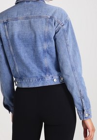 Veste en denim bleu clair avec un design court. Dotée de boutons à la taille et d'un col classique. Tissu texturé avec des coutures contrastantes.