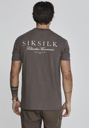 Brūns kokvilnas T-krekls ar "SIKSILK" un "Follow the Movement" baltā krāsā izdrukātu uz muguras. Īsās piedurknes un apaļa kakla izgriezuma forma.