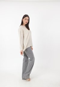 Maglione oversized in maglia beige con scollatura rotonda, abbinato a jeans grigi chiari a zampa d'elefante e sandali chiari, su uno sfondo bianco.