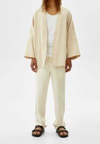 Person med lös beige kimono-jacka, matchande byxor, vit linne och svarta sandaler som står mot vit bakgrund.
