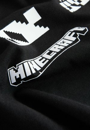 Tessuto di cotone nero con logo bianco di Minecraft e modelli grafici a blocchi. Il materiale è morbido con una texture liscia, leggermente piegato.