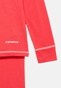 Prim-plan al unei cămăși termice roșii cu mânecă lungă, cu cusături albe și logo-ul alb Icepeak aproape de tiv, pe un fundal deschis.