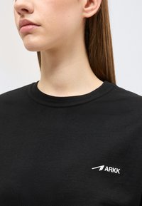 Γυναίκα με ίσια μαλλιά που φοράει μαύρο t-shirt ARKK με στρογγυλή λαιμόκοψη, ορατό από το λαιμό έως το στήθος.