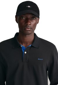 Schwarzes Langarm-Polohemd mit blauer Kragen und GANT-Logo. Das Modell trägt eine schwarze Cordmütze. Glatte Textur, klassisches Design und Knopfdetail.