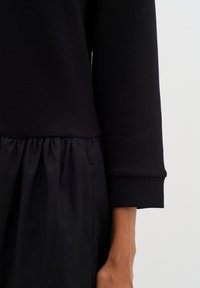 Schwarzes Kleid mit einem langärmeligen Oberteil und einem taillierten Oberkörper sowie einem gerafften, strukturierten Rock.Weiches Gewebe mit einer glatten Oberfläche.