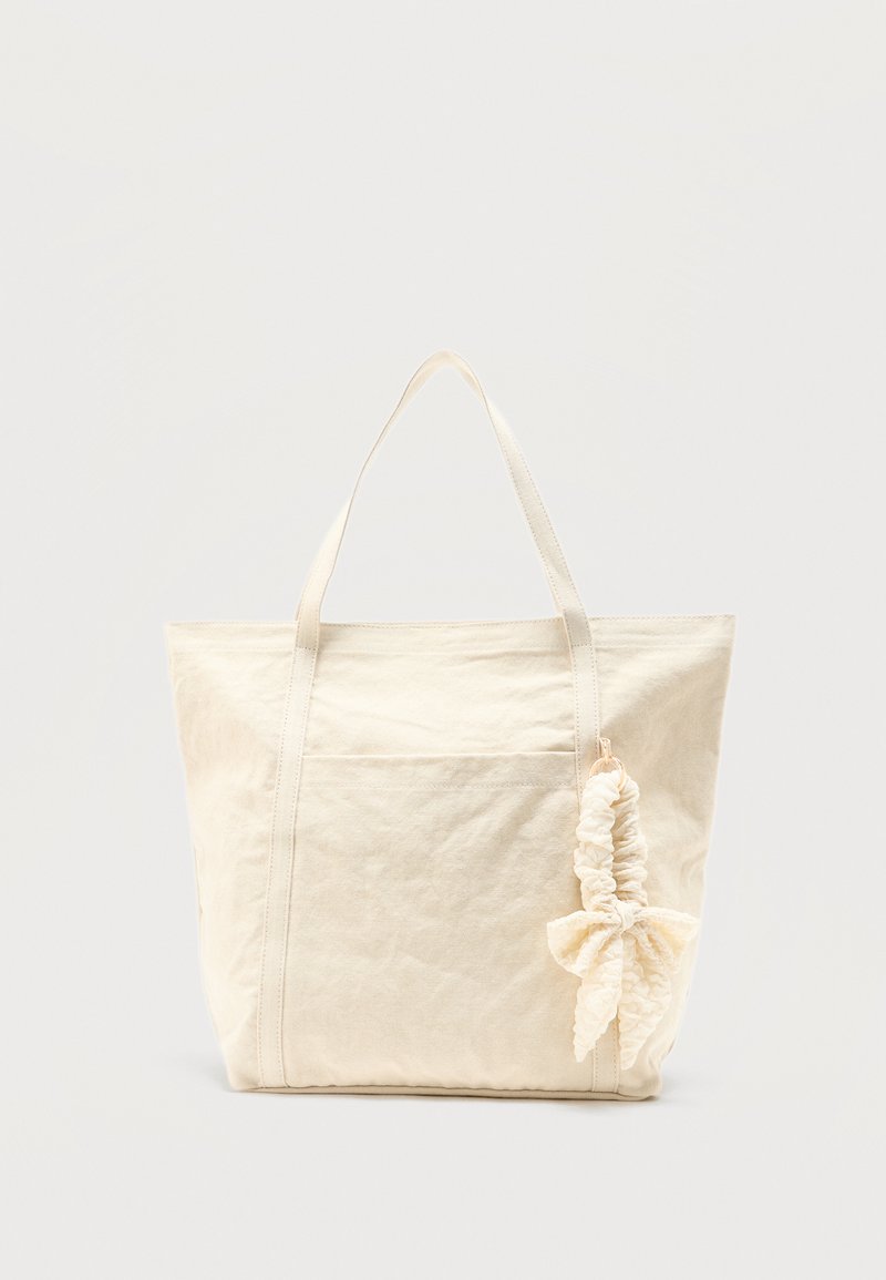 Sac cabas en toile beige avec deux poignées et un chouchou avec un nœud attaché à une poignée, sur un fond uni.
