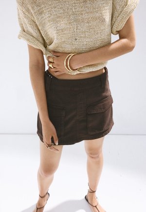 Femme portant un top court en maille beige, une minijupe marron foncé avec des poches, des bracelets et des bagues en or, et des sandales noires à lanières, debout sur un sol blanc.