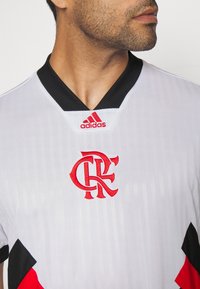 Maillot Adidas blanc avec col noir et accents rouges. Présente le logo "CRF" en broderie rouge sur la poitrine, avec des rayures verticales et des panneaux latéraux noirs et rouges.