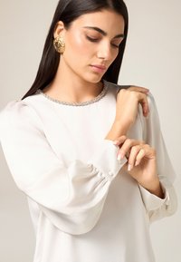 Langärmlige cremefarbene Bluse mit strukturiertem, schimmerndem Finish, verziert mit einem perlenbesetzten Ausschnitt und Knopfakzenten an den Manschetten.