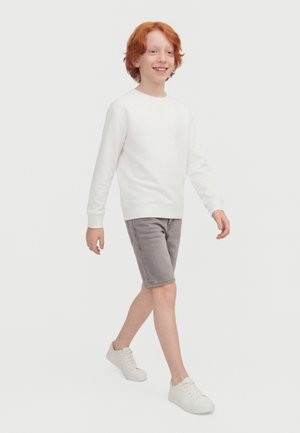 Smilende rødhåret dreng går, iført en hvid langærmet skjorte, grå shorts og hvide sneakers mod en ensfarvet baggrund.