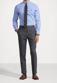 Man som bär en ljusblå skjorta, marinrandig slips, grå kostymbyxor, bruna loafers, blåmönstrade strumpor och en armbandsur.