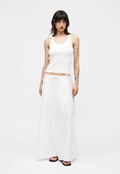 Γυναίκα με σκούρα μαλλιά μέχρι τους ώμους, φορά λευκό αμάνικο τοπ, μακριά λευκή φούστα, μαύρα σανδάλια και ορατά τατουάζ στα χέρια.