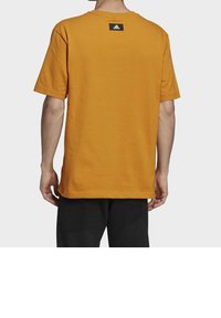 Orange bomull t-shirt med korta ärmar, rund halsringning, och en svart logotypetikett på övre ryggen. Bärs med svarta byxor.