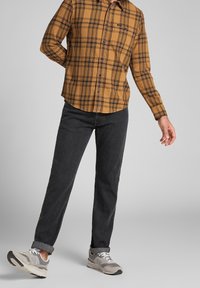 Camicia di flanella a scacchi marrone e nera, jeans neri e sneaker grigie con lacci bianchi. La camicia presenta una chiusura con bottoni e un orlo arrotondato.