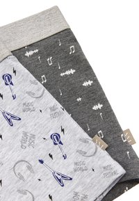 Dos pares de calcetines: uno gris claro con patrones de guitarras eléctricas y notas musicales, y el otro gris oscuro con ondas sonoras y símbolos musicales.