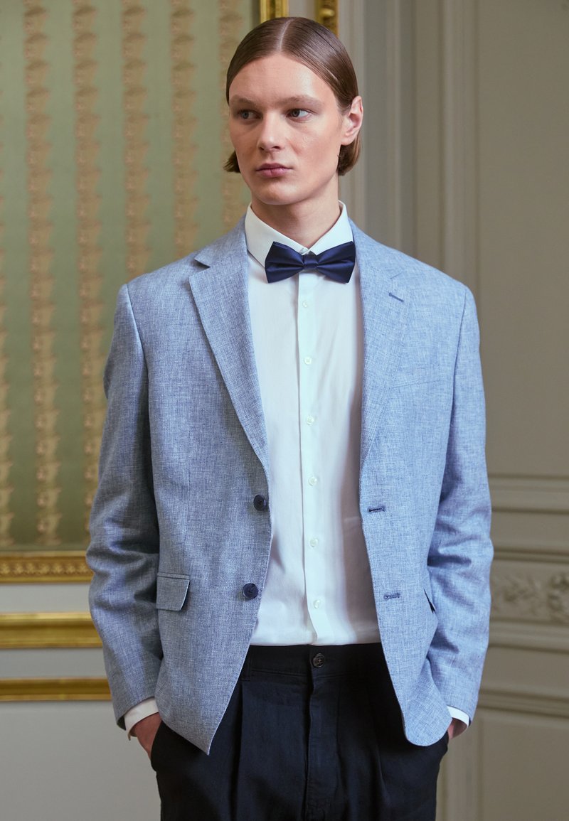 Gai zils lina žakets ar divām pogām, izgrieztiem atlaižām un priekšējiem kabatām, apvienots ar baltu krekli un tumši zilu tauriņu. Saskaņota tekstūra.