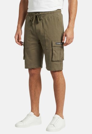 Homme portant un short cargo vert olive avec taille à cordon et poches plaquées, associé à des baskets blanches et un t-shirt blanc.